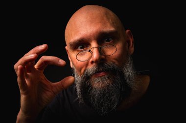 Gri sakallı ve yuvarlak gözlüklü gergin kel bir adam küçük bir çimdik hareketi yapıyor. Dramatik ışıklandırma ve siyah arkaplanda anlamlı yüz ile sade tuş portresi