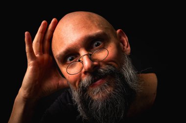 Gri sakallı ve yuvarlak gözlüklü gergin kel bir adam küçük bir çimdik hareketi yapıyor. Dramatik ışıklandırma ve siyah arkaplanda anlamlı yüz ile sade tuş portresi