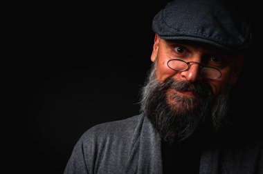 Gri şapkalı, gri kazaklı, siyah arka planda kameraya bakan duygusal sakallı bir adamın portresi. Loş ışıkta dramatik bir portre, etkileyici bir yüz, duygusal.