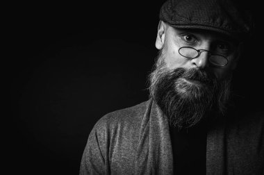 Siyah ve beyaz, gri şapkalı, gri kazaklı, siyah arka planda kameraya bakan duygusal sakallı bir adamın portresi. Loş ışıkta dramatik bir portre, etkileyici bir yüz, duygusal.