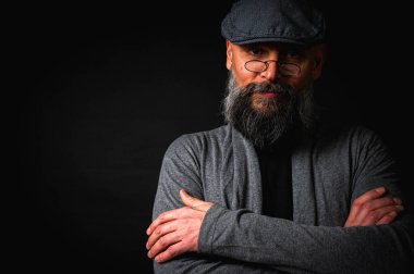 Gri şapkalı, gri kazaklı, siyah arka planda kameraya bakan duygusal sakallı bir adamın portresi. Loş ışıkta dramatik bir portre, etkileyici bir yüz, duygusal.