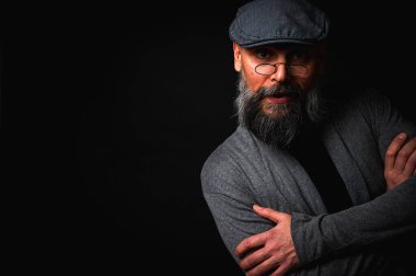 Gri şapkalı, gri kazaklı, siyah arka planda kameraya bakan duygusal sakallı bir adamın portresi. Loş ışıkta dramatik bir portre, etkileyici bir yüz, duygusal.