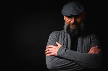 Gri şapkalı, gri kazaklı, siyah arka planda kameraya bakan duygusal sakallı bir adamın portresi. Loş ışıkta dramatik bir portre, etkileyici bir yüz, duygusal.