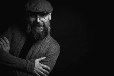 Siyah ve beyaz, gri şapkalı, gri kazaklı, siyah arka planda kameraya bakan duygusal sakallı bir adamın portresi. Loş ışıkta dramatik bir portre, etkileyici bir yüz, duygusal.