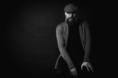 Siyah ve beyaz, gri şapkalı, gri kazaklı, siyah arka planda kameraya bakan duygusal sakallı bir adamın portresi. Loş ışıkta dramatik bir portre, etkileyici bir yüz, duygusal.