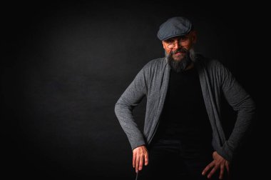Gri şapkalı, gri kazaklı, siyah arka planda kameraya bakan duygusal sakallı bir adamın portresi. Loş ışıkta dramatik bir portre, etkileyici bir yüz, duygusal.