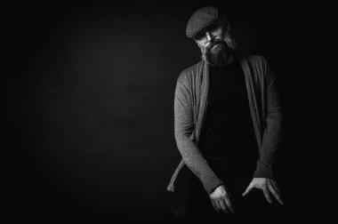 Siyah ve beyaz, gri şapkalı, gri kazaklı, siyah arka planda kameraya bakan duygusal sakallı bir adamın portresi. Loş ışıkta dramatik bir portre, etkileyici bir yüz, duygusal.