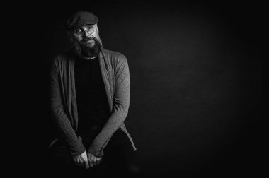 Siyah ve beyaz, gri şapkalı, gri kazaklı, siyah arka planda kameraya bakan duygusal sakallı bir adamın portresi. Loş ışıkta dramatik bir portre, etkileyici bir yüz, duygusal.