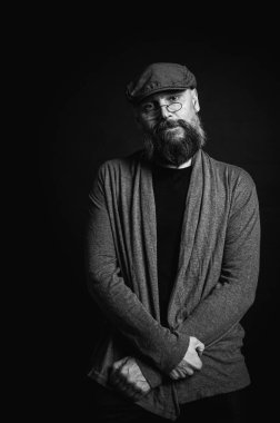 Siyah ve beyaz, gri şapkalı, gri kazaklı, siyah arka planda kameraya bakan duygusal sakallı bir adamın portresi. Loş ışıkta dramatik bir portre, etkileyici bir yüz, duygusal.