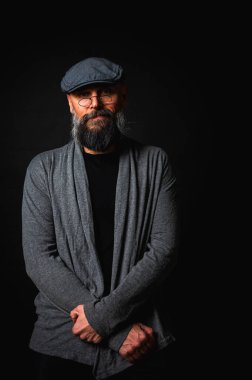 Gri şapkalı, gri kazaklı, siyah arka planda kameraya bakan duygusal sakallı bir adamın portresi. Loş ışıkta dramatik bir portre, etkileyici bir yüz, duygusal.