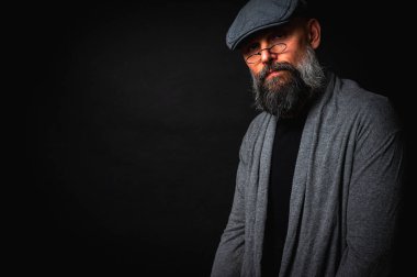 Gri pirinç muştalı, gri kazaklı ve siyah arka planda kameraya bakan duygusal sakallı bir adamın portresi. Bastırılmış aydınlatma, anlamlı yüz, duygusal gerilim, karanlık arka plan dramatik portre.