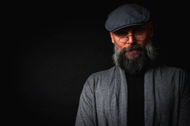 Düz şapkalı ve siyah arka planda gözlüklü ciddi bir sakallı adamın yakın plan portresi. Tarz sahibi, kendine güvenen ve gizemli ifadeli yoğun bakışlar.