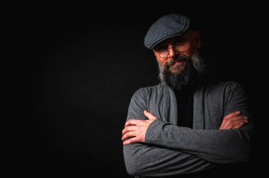 Düz şapkalı ve siyah arka planda gözlüklü ciddi bir sakallı adamın yakın plan portresi. Tarz sahibi, kendine güvenen ve gizemli ifadeli yoğun bakışlar.