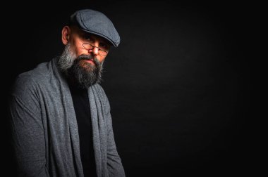 Düz şapkalı ve siyah arka planda gözlüklü ciddi bir sakallı adamın yakın plan portresi. Tarz sahibi, kendine güvenen ve gizemli ifadeli yoğun bakışlar.