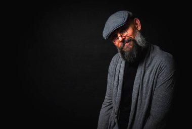 Düz şapkalı ve siyah arka planda gözlüklü ciddi bir sakallı adamın yakın plan portresi. Tarz sahibi, kendine güvenen ve gizemli ifadeli yoğun bakışlar.