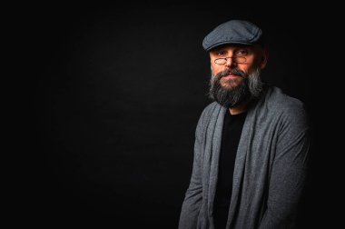 Düz şapkalı ve siyah arka planda gözlüklü ciddi bir sakallı adamın yakın plan portresi. Tarz sahibi, kendine güvenen ve gizemli ifadeli yoğun bakışlar.