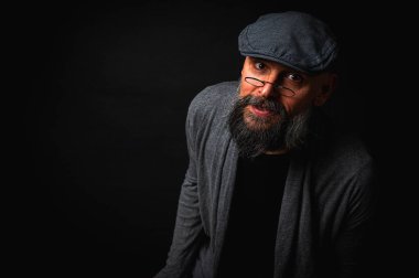 Ciddi sakallı, şapkalı ve siyah arka planda gözlüklü bir adamın duygusal, makro portresi. Şık, kendine güvenen ve gizemli bir ifade ile yoğun bir görünüm, bir reklam panosuna yazılacak duygular.