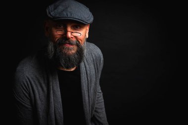 Düz şapkalı ve siyah arka planda gözlüklü ciddi bir sakallı adamın yakın plan portresi. Tarz sahibi, kendine güvenen ve gizemli ifadeli yoğun bakışlar.