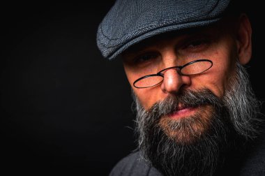 Düz şapkalı ve siyah arka planda gözlüklü ciddi bir sakallı adamın yakın plan portresi. Tarz sahibi, kendine güvenen ve gizemli ifadeli yoğun bakışlar.