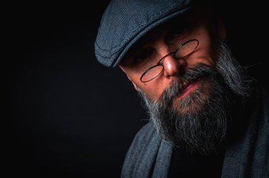 Düz şapkalı ve siyah arka planda gözlüklü ciddi bir sakallı adamın yakın plan portresi. Tarz sahibi, kendine güvenen ve gizemli ifadeli yoğun bakışlar.