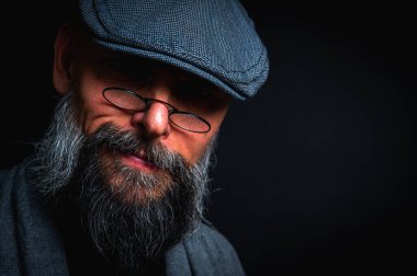 Düz şapkalı ve siyah arka planda gözlüklü ciddi bir sakallı adamın yakın plan portresi. Tarz sahibi, kendine güvenen ve gizemli ifadeli yoğun bakışlar.