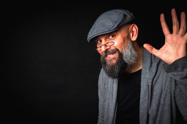 Şapkalı ve gözlüklü yaşlı sakallı bir adam el hareketi yapıyor, alaycılık ya da başkaldırı gösteriyor. Siyah arkaplanda stüdyo resmi