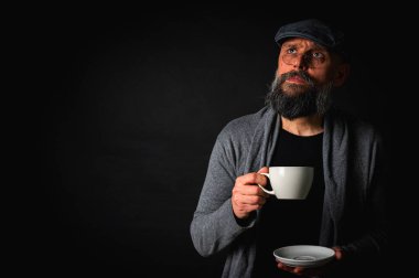 Siyah arka planda kahve fincanı ve fincan tabağı tutan şapkalı şık sakallı bir adam. Düşünceli ifade, rahat giysi, modern klasik atmosfer, sabah çayı.