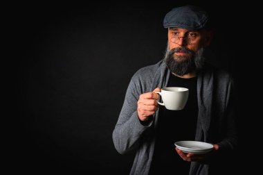 Siyah arka planda kahve fincanı ve fincan tabağı tutan şapkalı şık sakallı bir adam. Düşünceli ifade, rahat giysi, modern klasik atmosfer, sabah çayı.