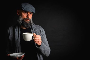 Siyah arka planda kahve fincanı ve fincan tabağı tutan şapkalı şık sakallı bir adam. Düşünceli ifade, rahat giysi, modern klasik atmosfer, sabah çayı.