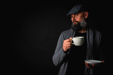 Siyah arka planda kahve fincanı ve fincan tabağı tutan şapkalı şık sakallı bir adam. Düşünceli ifade, rahat giysi, modern klasik atmosfer, sabah çayı.