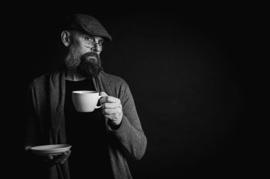 Siyah beyaz fotoğraf, şapkalı şık sakallı adam elinde kahve fincanı ve arka planda fincan tabağı tutan gözlüklü adam. Düşünceli ifade, rahat rahat kıyafetler, modern klasik atmosfer, sabah çay partisi.
