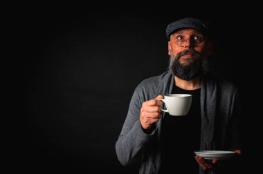 Siyah arka planda kahve fincanı ve fincan tabağı tutan şapkalı şık sakallı bir adam. Düşünceli ifade, rahat giysi, modern klasik atmosfer, sabah çayı.