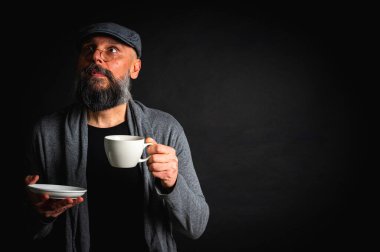 Siyah arka planda kahve fincanı ve fincan tabağı tutan şapkalı şık sakallı bir adam. Düşünceli ifade, rahat giysi, modern klasik atmosfer, sabah çayı.