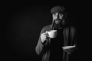 Siyah beyaz fotoğraf, şapkalı şık sakallı adam elinde kahve fincanı ve arka planda fincan tabağı tutan gözlüklü adam. Düşünceli ifade, rahat rahat kıyafetler, modern klasik atmosfer, sabah çay partisi.
