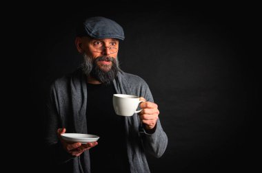 Siyah arka planda kahve fincanı ve fincan tabağı tutan şapkalı şık sakallı bir adam. Düşünceli ifade, rahat giysi, modern klasik atmosfer, sabah çayı.