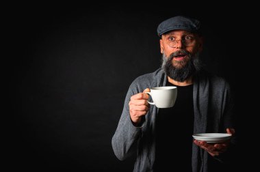 Siyah arka planda kahve fincanı ve fincan tabağı tutan şapkalı şık sakallı bir adam. Düşünceli ifade, rahat giysi, modern klasik atmosfer, sabah çayı.