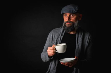 Siyah arka planda kahve fincanı ve fincan tabağı tutan şapkalı şık sakallı bir adam. Düşünceli ifade, rahat giysi, modern klasik atmosfer, sabah çayı.