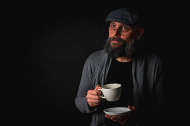 Siyah arka planda kahve fincanı ve fincan tabağı tutan şapkalı şık sakallı bir adam. Düşünceli ifade, rahat giysi, modern klasik atmosfer, sabah çayı.