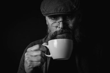 Siyah beyaz fotoğraf, şapkalı şık sakallı adam elinde kahve fincanı ve arka planda fincan tabağı tutan gözlüklü adam. Düşünceli ifade, rahat rahat kıyafetler, modern klasik atmosfer, sabah çay partisi.