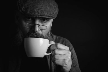 Siyah beyaz fotoğraf, şapkalı şık sakallı adam elinde kahve fincanı ve arka planda fincan tabağı tutan gözlüklü adam. Düşünceli ifade, rahat rahat kıyafetler, modern klasik atmosfer, sabah çay partisi.