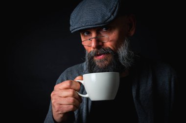 Siyah arka planda kahve fincanı ve fincan tabağı tutan şapkalı şık sakallı bir adam. Düşünceli ifade, rahat giysi, modern klasik atmosfer, sabah çayı.