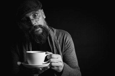 Siyah beyaz fotoğraf, şapkalı şık sakallı adam elinde kahve fincanı ve arka planda fincan tabağı tutan gözlüklü adam. Düşünceli ifade, rahat rahat kıyafetler, modern klasik atmosfer, sabah çay partisi.