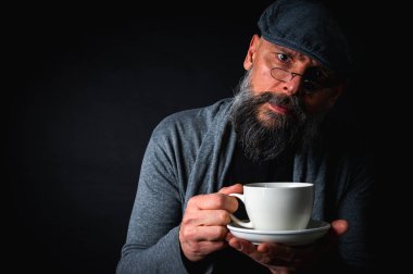 Siyah arka planda kahve fincanı ve fincan tabağı tutan şapkalı şık sakallı bir adam. Düşünceli ifade, rahat giysi, modern klasik atmosfer, sabah çayı.