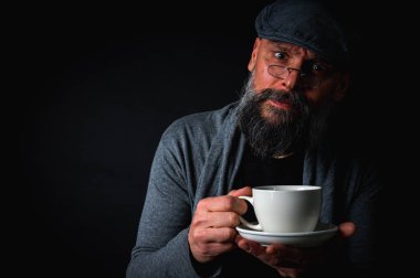 Siyah arka planda kahve fincanı ve fincan tabağı tutan şapkalı şık sakallı bir adam. Düşünceli ifade, rahat giysi, modern klasik atmosfer, sabah çayı.