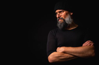 Gözlüklü, düz şapkalı, gölgede oturan düşünceli sakallı adam. Kollarını arkada kavuşturdu. Derin kontrastlı ve karamsar ışıklandırmalı stüdyo portresi