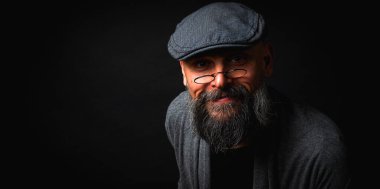 Düz şapkalı, gözlüklü ve kaşlarını kaldırmış gülümseyen kendine güvenen sakallı bir adam. Siyah arkaplanı olan bir stüdyo resmi. Zeki, çekici ve şık olgun bir erkek.