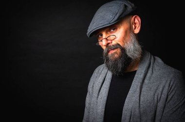 Düz şapkalı, gözlüklü ve kaşlarını kaldırmış gülümseyen kendine güvenen sakallı bir adam. Siyah arkaplanı olan bir stüdyo resmi. Zeki, çekici ve şık olgun bir erkek.