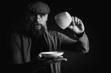 Siyah ve beyaz portre, şık şapkalı ve gözlüklü olgun sakallı bir adam gizemli bir gülümsemeyle bir fincan kahve sunuyor. Olağanüstü aydınlatma ve karanlık arka plan eski moda bir hava yaratıyor..