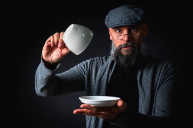 Boş bir fincan ve fincan tabağı tutan düz şapkalı sakallı adam şaşırdı, kaşlarını kaldırarak kontrol ediyor. Komik ifade, karanlık arka planlı stüdyo çekimi.