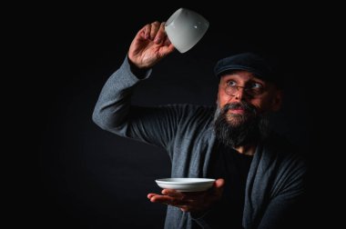 Boş bir fincan ve fincan tabağı tutan düz şapkalı sakallı adam şaşırdı, kaşlarını kaldırarak kontrol ediyor. Komik ifade, karanlık arka planlı stüdyo çekimi.